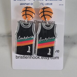 May include: A pair of earrings featuring orange basketball studs attached to black basketball jersey dangles. The jerseys display the text San Antonio, a red, orange, and yellow stripe, and the number 1. The display card reads Bristle 'n' Hook Creations.