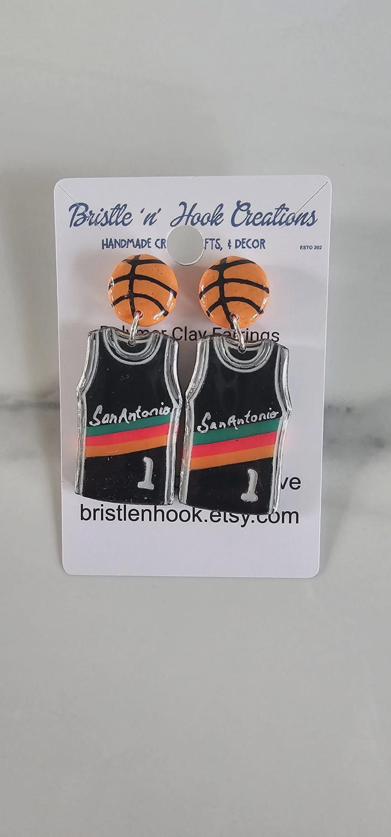 May include: A pair of earrings featuring orange basketball studs attached to black basketball jersey dangles. The jerseys display the text San Antonio, a red, orange, and yellow stripe, and the number 1. The display card reads Bristle 'n' Hook Creations.