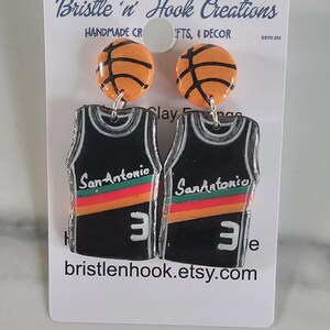 May include: A pair of dangle earrings featuring orange basketball studs and black jersey-shaped pendants. The jerseys display the text San Antonio, a red and orange horizontal stripe, and the number 3 in white. The display card reads Bristle 'n Hook Creations.