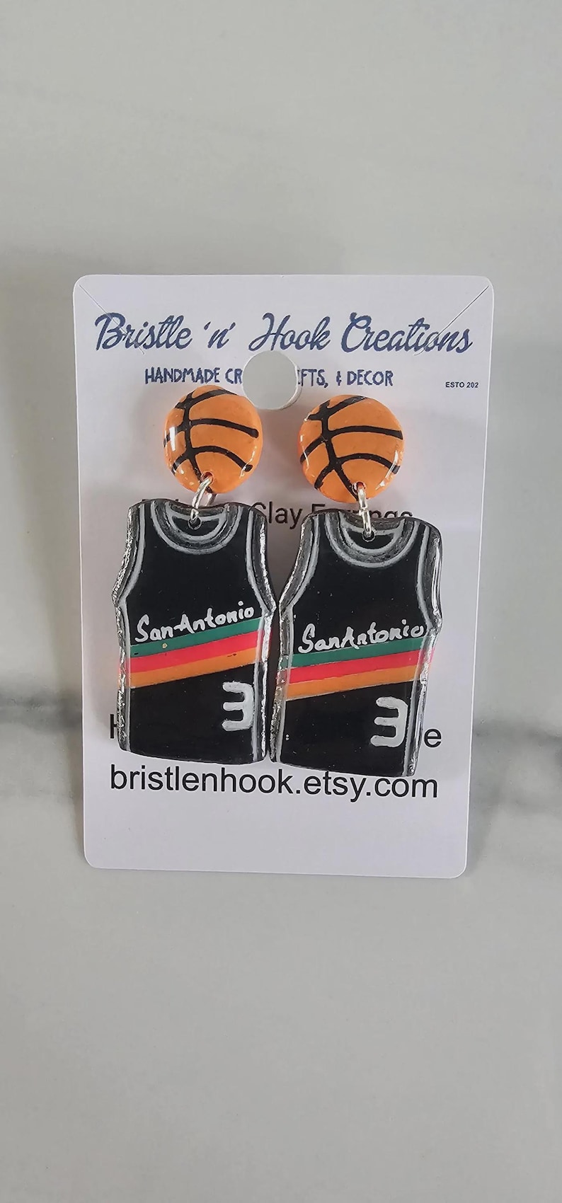 May include: A pair of dangle earrings featuring orange basketball studs and black jersey-shaped pendants. The jerseys display the text San Antonio, a red and orange horizontal stripe, and the number 3 in white. The display card reads Bristle 'n Hook Creations.