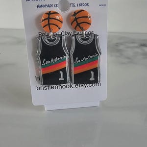 May include: A pair of earrings on a display card featuring orange basketball studs and black jersey-shaped dangles with the text San Antonio, a red and orange stripe, and the number 1. The card displays the text Bristle 'n' Hook Creations.
