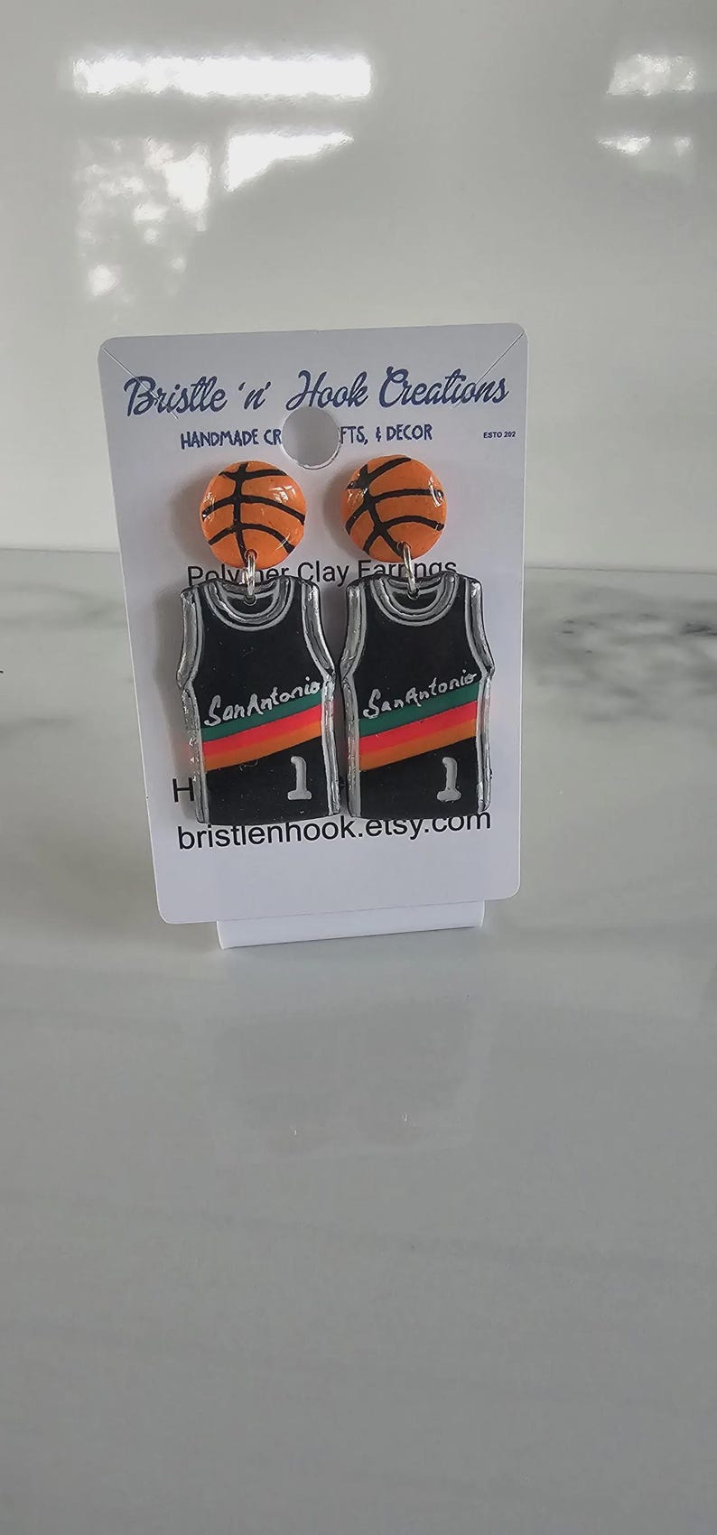 May include: A pair of earrings on a display card featuring orange basketball studs and black jersey-shaped dangles with the text San Antonio, a red and orange stripe, and the number 1. The card displays the text Bristle 'n' Hook Creations.