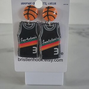 May include: A pair of dangle earrings featuring orange basketball studs and black jersey pendants. The jerseys display the text San Antonio, a red, orange, and yellow stripe, and the number 3 in white. The display card reads Bristle 'n' Hook Creations.