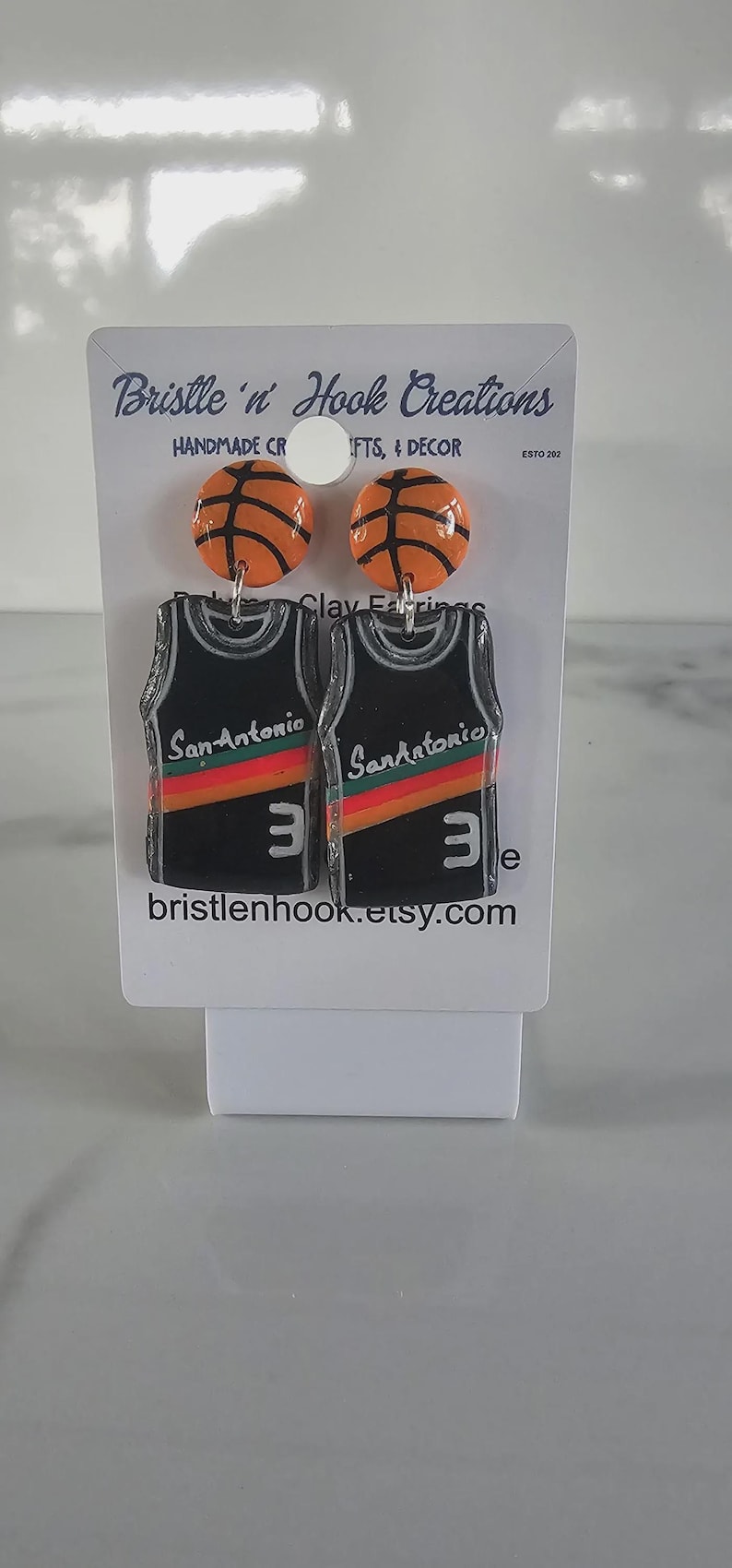 May include: A pair of dangle earrings featuring orange basketball studs and black jersey pendants. The jerseys display the text San Antonio, a red, orange, and yellow stripe, and the number 3 in white. The display card reads Bristle 'n' Hook Creations.