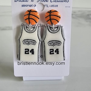 Pode incluir: Um par de brincos num cartão branco com tachas de basquetebol laranja e pendentes de camisola de basquetebol brancos com rebordo preto, o logótipo dos Spurs e o número 24. O cartão apresenta o texto Bristle 'n Hook Creations e bristlennook.etsy.com.