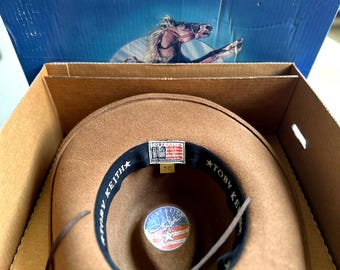 May include: A brown cowboy hat with a leather chin strap sits inside a cardboard box. The hat has a black interior band with "TOBY KEITH" printed on it. The box has the word "RESISTOL" printed on it.