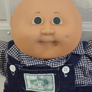 May include: A Cabbage Patch Kids doll with a round, bald head, light skin, and green eyes. The doll wears a navy blue denim overall with white buttons over a black and white gingham shirt. A rectangular label on the chest reads Cabbage Patch Kids.