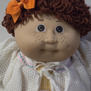 May include: A doll with brown yarn hair styled in curls, featuring an orange ribbon bow on the side. The doll has a round face with freckles, dark eyes, and a neutral expression. It wears a white knit cardigan over a yellow and white dress.
