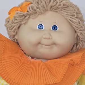 Puede incluir: Una muñeca con rostro redondo de tono claro, ojos azul brillante y una expresión suave y sonriente. La muñeca tiene cabello de lana marrón claro con un pequeño lazo naranja y lleva un cuello de tela plisado grande de color naranja.