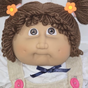 Könnte beinhalten: Eine Puppe mit braunem Wollhaar, das zu zwei Zöpfen gebunden ist, die jeweils mit einer orangefarbenen Blumenklammer verziert sind. Die Puppe hat braune Augen, Grübchen und trägt ein weißes Hemd unter einer beigen Cordlatzhose mit rosa Knöpfen.