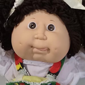 Könnte beinhalten: Eine Puppe mit einem weichen Vinylgesicht, braunen Augen und Grübchen. Die Puppe hat dunkelbraunes Wollhaar, das zu zwei Zöpfen mit weißen Bändern frisiert ist. Sie trägt ein weißes Hemd unter einem Latzkleid aus grünem, rotem, gelbem und blauem Cord.