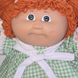 May include: A doll with orange yarn hair styled in two pigtails with white ribbons. The doll has green eyes, a round face with a dimple, and wears a green and white gingham dress featuring a white collar with eyelet lace and a white tie.