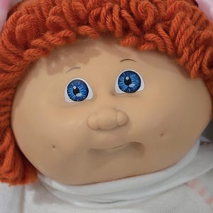 May include: A doll with a soft, light-toned vinyl face, bright blue eyes, and a small nose. The doll features thick, textured orange yarn hair styled in a short cut and wears a plain white fabric collar around the neck area.