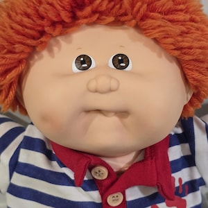 May include: A doll with a round face, brown eyes, and textured orange yarn hair. The doll wears a white and blue horizontal striped shirt with a red collar, two light-colored buttons, and a red handprint graphic with the letters CPK.