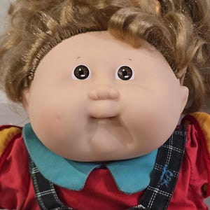May include: A doll with light skin, brown eyes, and curly brown hair tied in a ponytail. The doll wears a red shirt with a teal collar under black overalls featuring a white plaid pattern, yellow buttons, and the text CPK printed on the fabric.