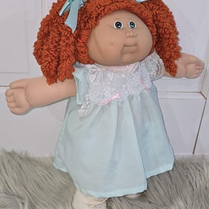 May include: A doll with curly auburn yarn hair styled in pigtails with light blue satin ribbons. The doll has a round face, blue eyes, and wears a light blue dress featuring a white lace collar with small pink ribbon accents and white shoes.