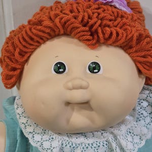 May include: A doll with a round, light-toned face, green eyes, and textured orange yarn hair styled with a purple fabric bow. The doll wears a light blue garment featuring a white lace collar with a delicate floral pattern along the neckline.