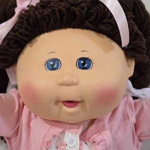 May include: A doll with a round face, blue eyes, and rosy cheeks, featuring dark brown yarn hair styled in pigtails with light pink ribbons. The doll wears a light pink cardigan over a white shirt with a pink floral pattern and a single white button.