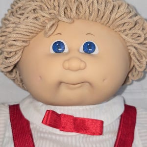 May include: A doll with light tan skin, bright blue eyes, and dimpled cheeks. The doll has textured beige yarn hair styled in a short cut. It wears a white ribbed shirt with a small red bow at the collar and red corduroy overall straps.