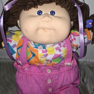 May include: A doll with brown yarn hair styled in pigtails with light purple ribbons and a small blue butterfly clip. The doll has blue eyes, a soft vinyl face, and wears a white shirt with a colourful geometric pattern under pink overalls.