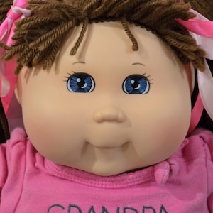 Può includere: Una bambola con viso rotondo, occhi blu e capelli di lana marrone acconciati in codini con nastri rosa. La bambola indossa una maglietta rosa con la scritta "GRANDPA LOVES" ricamata in grigio sul davanti. Ha un incarnato chiaro e liscio.