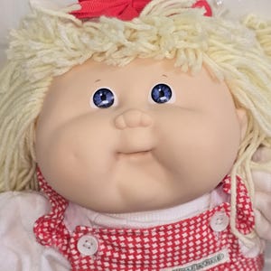 May include: A doll with pale skin, blue eyes, and blonde yarn hair styled with a red bow. The doll wears a white shirt under red and white checkered overalls featuring two white buttons and a small label that reads Cabbage Patch Kids.