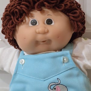 May include: A doll with brown yarn hair, a round face, and dimples, wearing a light blue ribbed overall outfit over a white shirt. The overalls feature a small embroidered gray elephant with pink ears and feet on the front chest panel.
