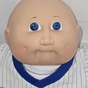May include: A bald doll with a light skin tone, blue eyes, and dimpled cheeks. The doll wears a white shirt with thin blue pinstripes, a blue V-neck collar, and the word BREWERS printed in blue capital letters across the chest area.