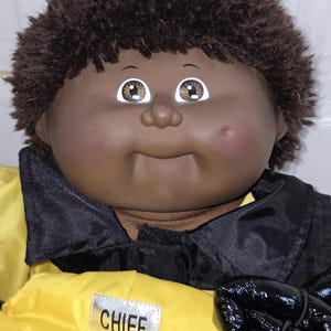 Puede incluir: Una muñeca de piel marrón oscuro, ojos marrones y cabello corto y rizado de color marrón oscuro. La muñeca viste una chaqueta amarilla y negra con un parche plateado en el pecho que tiene la palabra CHIEF en letras mayúsculas.