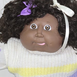 May include: A doll with dark brown curly hair, brown eyes, and a round face. The doll features a purple ribbon on the left side and a white ribbon on the right side of the hair. It wears a sleeveless white knit sweater with yellow and purple stripes.