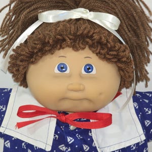 May include: A doll with brown yarn hair styled with a white satin ribbon, blue eyes, and a dimpled face. The doll wears a blue shirt featuring a white nautical print of sailboats and anchors, accented by a white sailor collar and a red ribbon tie.