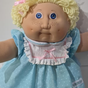 Könnte beinhalten: Eine Puppe mit lockigem gelbem Wollhaar, blauen Augen und Sommersprossen auf den Wangen. Sie trägt ein hellblaues Kleid mit weißen Punkten, einen weißen Spitzenkragen mit rosa Schleifen und ein kleines weißes Etikett unten rechts am Kleid.