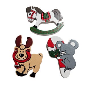 May include: Three wooden Christmas decorations: a rocking horse, a reindeer, and a mouse holding a candy cane. The horse is white with red and green details, the reindeer is brown with red and white details, and the mouse is grey with red and green details.