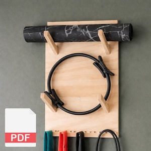 Op de afbeelding: Een houten wandorganizer voor fitnessapparatuur. Het heeft een opgerolde zwarte yogamat met witte marmering, een zwarte Pilates-ring en verschillende gekleurde weerstandsbanden. De organizer is gemaakt van lichtgekleurd hout.