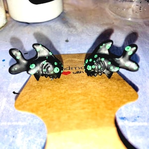 May include: A pair of black shark-shaped earrings with green polka dots and accents. The earrings are displayed on a brown card with the text "handmade with" and a small red heart.