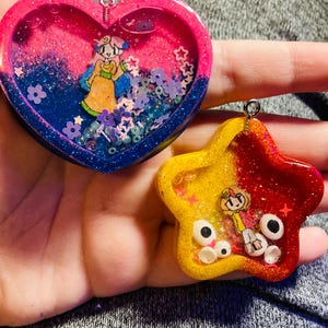 May include: Two handmade resin charms. One is heart-shaped with pink and blue glitter, a cartoon figure, and flower accents. The other is star-shaped, yellow and red glitter, with a cartoon figure and white accents.