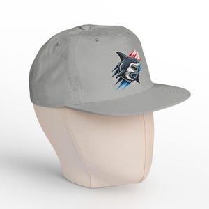 May include: A light gray baseball cap featuring a graphic of a shark with bared teeth, accented by red and blue motion lines. The cap has a structured crown, a flat brim, and eyelets for ventilation, presenting a clean and simple design.