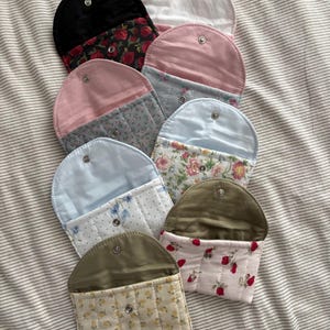 May include: A collection of handmade fabric pouches in various colors and patterns. The pouches feature a snap closure and are made from materials like velvet, floral prints, and quilted fabrics. Colors include black, pink, blue, and white.