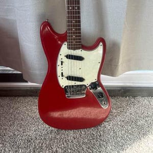 May include: A red electric guitar with a white pickguard and chrome hardware. The guitar has a classic design with a glossy finish. The strings and frets are visible on the neck. The guitar is resting on a gray carpet.