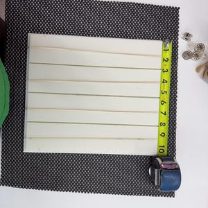 Può includere: Una tavola rettangolare bianca con sette fessure orizzontali, posizionata accanto a un metro a nastro giallo che indica un'altezza di circa 25,4 cm. La tavola ha una superficie liscia ed è appoggiata su un tappetino nero testurizzato.