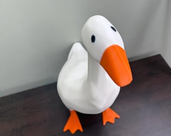 May include: A white goose figurine with an orange beak and feet. The goose has black eyes and is positioned on a dark brown surface. The figurine is a decorative item.