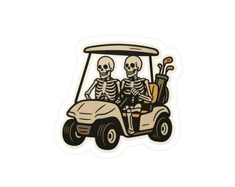 May include: A sticker featuring two skeleton figures in a golf cart. The cart is beige with black wheels, and a golf bag with clubs is visible. The skeletons are in the driver and passenger seats.