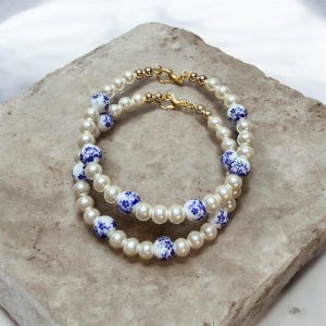 Może przedstawiać: Dwie perłowe bransoletki ze złotymi zapięciami i niebiesko-białymi kwiatowymi ceramicznymi koralikami. Bransoletki ułożone są na jasnej kamiennej powierzchni. Perły są okrągłe i równomiernej wielkości.