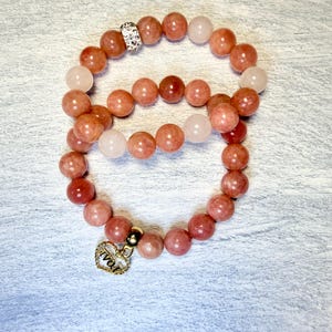 Może przedstawiać: Dwie bransoletki z koralików. Jedna bransoletka ma brzoskwiniowe koraliki i srebrny separator. Druga bransoletka ma brzoskwiniowe i jasnoróżowe koraliki, ze złotym sercem z napisem "love".