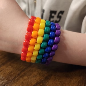 May include: A rainbow-colored beaded bracelet. The bracelet features rows of red, orange, yellow, green, blue, and purple beads. The bracelet is worn on a wrist.