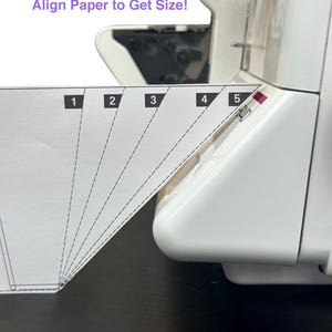 Może przedstawiać: Biała maszyna do szycia z prowadnicą papieru oznaczoną od 1 do 5. Tekst "Dopasuj papier, aby uzyskać rozmiar!" znajduje się nad prowadnicą. Maszyna stoi na czarnej powierzchni.