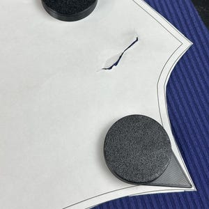 Może przedstawiać: Biały papierowy wz&oacute;r z rozerwanym fragmentem, utrzymywany na miejscu przez dwa czarne, okrągłe magnesy. Wz&oacute;r umieszczony jest na niebieskiej, prążkowanej powierzchni materiału. Magnesy mają teksturowaną powierzchnię.