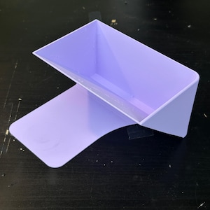 Może przedstawiać: Lawendowa p&oacute;łka wydrukowana w technologii 3D z prostokątnym miejscem do przechowywania i zakrzywionym wspornikiem. P&oacute;łka jest przeznaczona do montażu na ścianie lub umieszczenia na płaskiej powierzchni. Projekt jest minimalistyczny i nowoczesny.