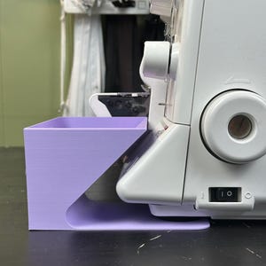 Może przedstawiać: Biała maszyna do szycia z fioletowym akcesorium przymocowanym z boku. Akcesorium to mały pojemnik z otwartą g&oacute;rą. Maszyna posiada włącznik/wyłącznik i duże, okrągłe koło. Tło to jasnozielona ściana.