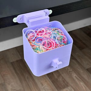 Può includere: Un contenitore di stoccaggio lilla stampato in 3D pieno di elastici colorati. Il contenitore è progettato per essere appeso a una scrivania o a uno scaffale. Ha una maniglia e una staffa di montaggio.